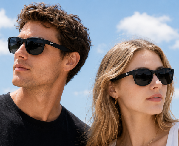 A man and a woman wearing black sunglasses with a small white logo on the temple, photographed outdoors against a bright blue sky with clouds.