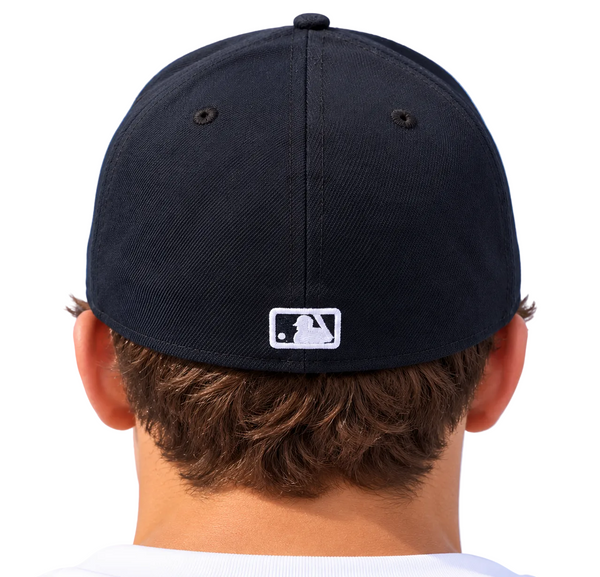 Rear view of a person wearing a dark navy baseball cap with a white MLB logo patch on the back, set against a light blue sky.
